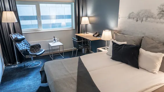 Säng prydligt bäddad med grått och vitt sänglinne; belägen i ett modernt hotellrum med en svart stol, litet bord med vin, skrivbord och mjuk belysning. Stort fönster ger gott om naturligt ljus.