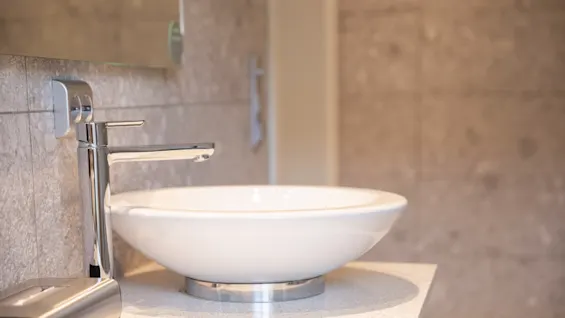 A white, round sink sits on a countertop, next to a sleek, chrome faucet in a modern bathroom with gray stone walls.