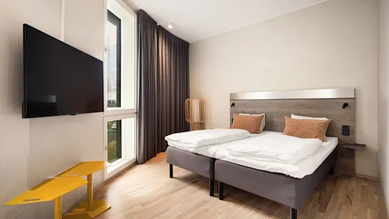 A neatly made bed with white and brown pillows stands in a bright room with light wooden floors, a wall-mounted television, and large window with dark curtains.