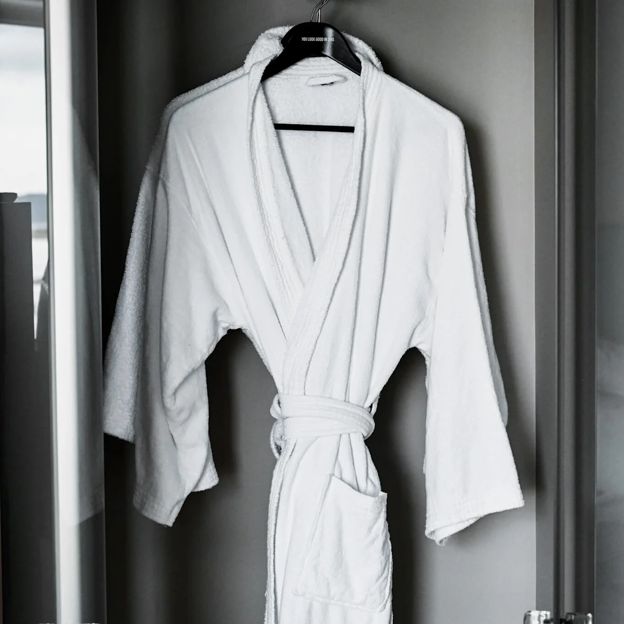 White bathrobe hanging on a black hanger inside a glass-doored wardrobe. Text on the hanger reads: "YOU LOOK GOOD IN ME". 