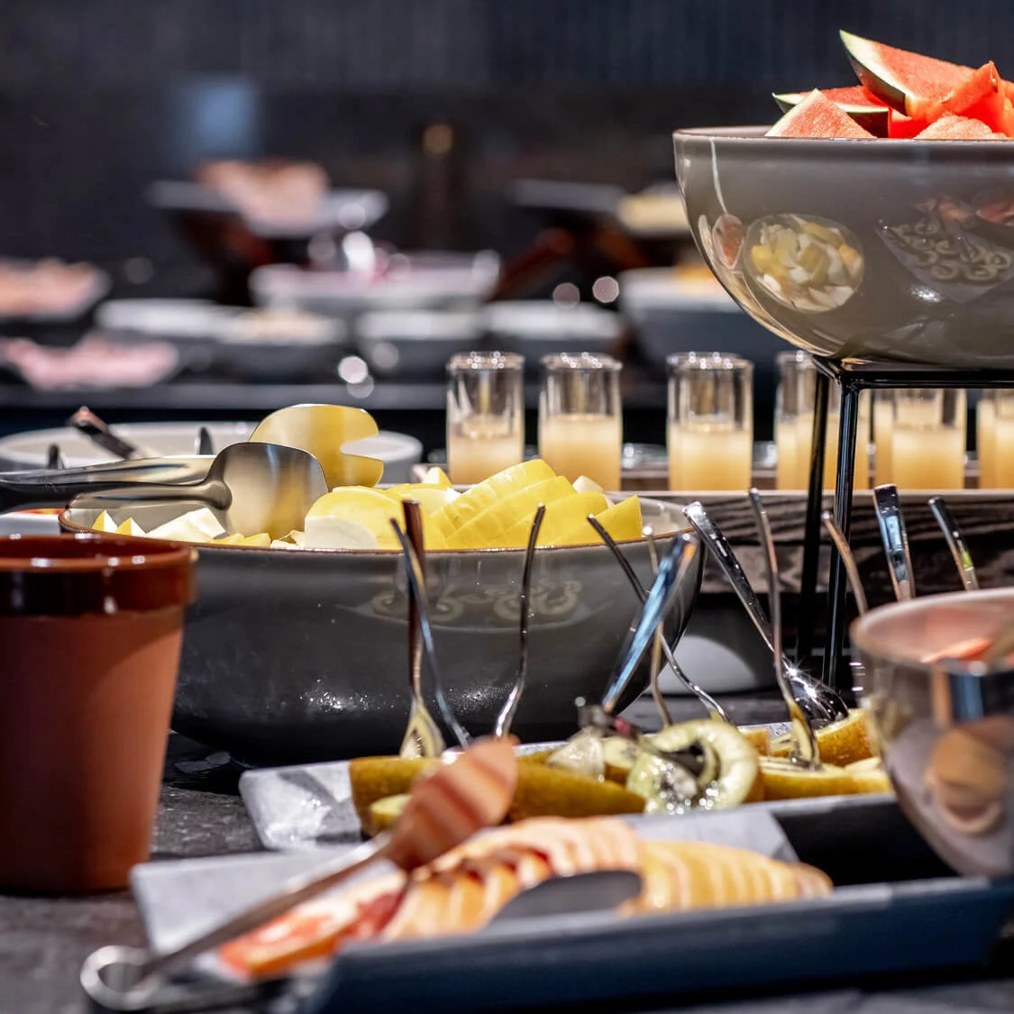 Skåler med frugt, herunder vandmelon og ananas, vises på et buffetbord sammen med juiceglas. Forskellige redskaber og tallerkener skaber en livlig, indbydende spisestemning.