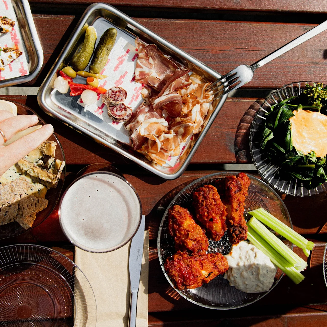 A hand reaches for sliced bread beside plates of pickles, charcuterie, wings, dip, and a leafy dish topped with a fried egg on an outdoor wooden table. 