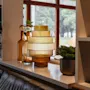 A cozy indoor scene featuring a tiered amber glass lamp, decorative bottles, and potted plants by a large window overlooking lush greenery.