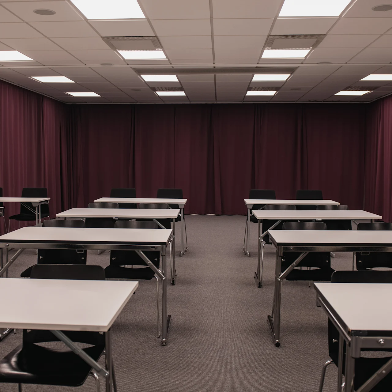 Tables and chairs are arranged in rows facing forward in a classroom with purple curtains and a ceiling of bright lights.