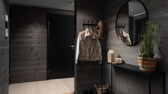 A hallway features a coat rack with jackets and a hat. A round mirror hangs above a black console table holding a plant in a basket, near a dark wooden door.