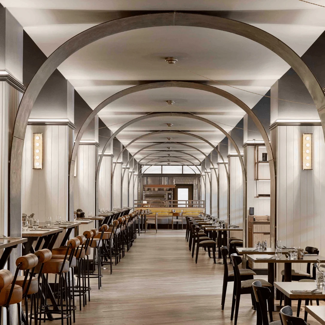 Restaurant interior features rows of wooden tables and chairs lined with elegant arches, under soft, warm lighting. The symmetrical design creates a cozy and inviting dining atmosphere.