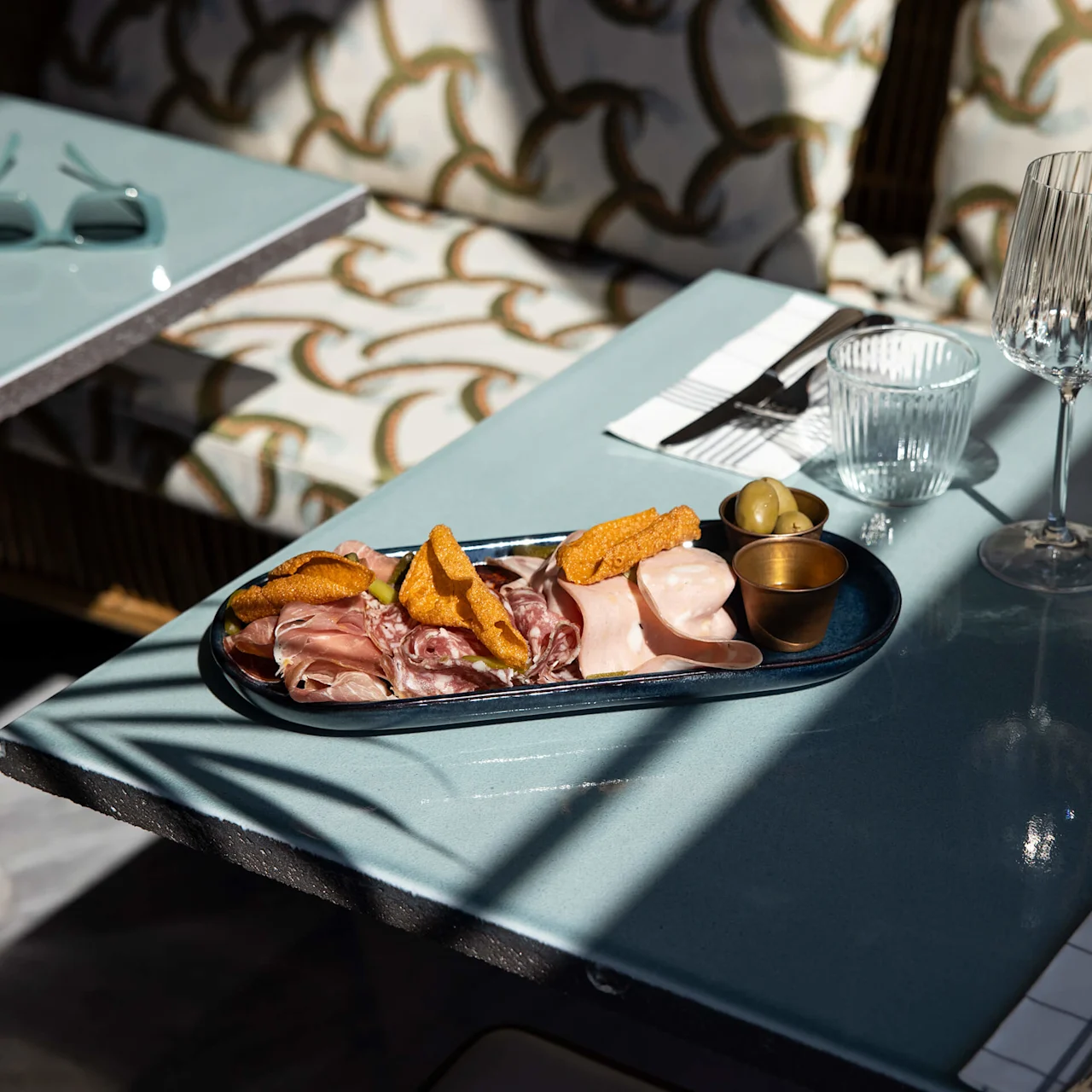 A charcuterie platter with cold cuts, crisps, and olives sits on a light blue table. Nearby, empty glasses and silverware are neatly arranged under dappled sunlight with patterned cushions.