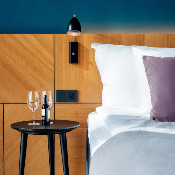 A cozy hotel room with a bedside table holding wine glasses and a bottle, next to a bed with a purple cushion.