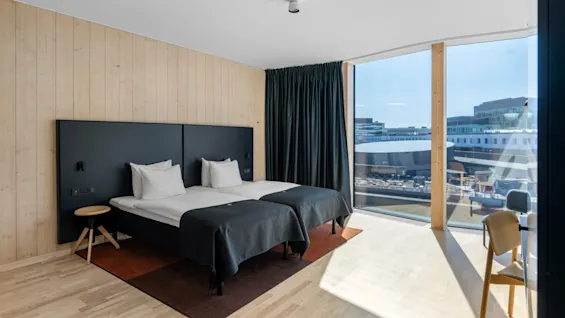 Et moderne hotelværelse med to senge, trævægge og et stort vindue med udsigt over et bybillede.