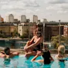 A group of people relax in a rooftop pool overlooking a cityscape with historical buildings. Visible text: "#POOLCLUB #SELMACITYSPA STHLM".