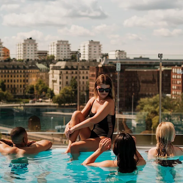 A group of people relax in a rooftop pool overlooking a cityscape with historical buildings. Visible text: "#POOLCLUB #SELMACITYSPA STHLM".
