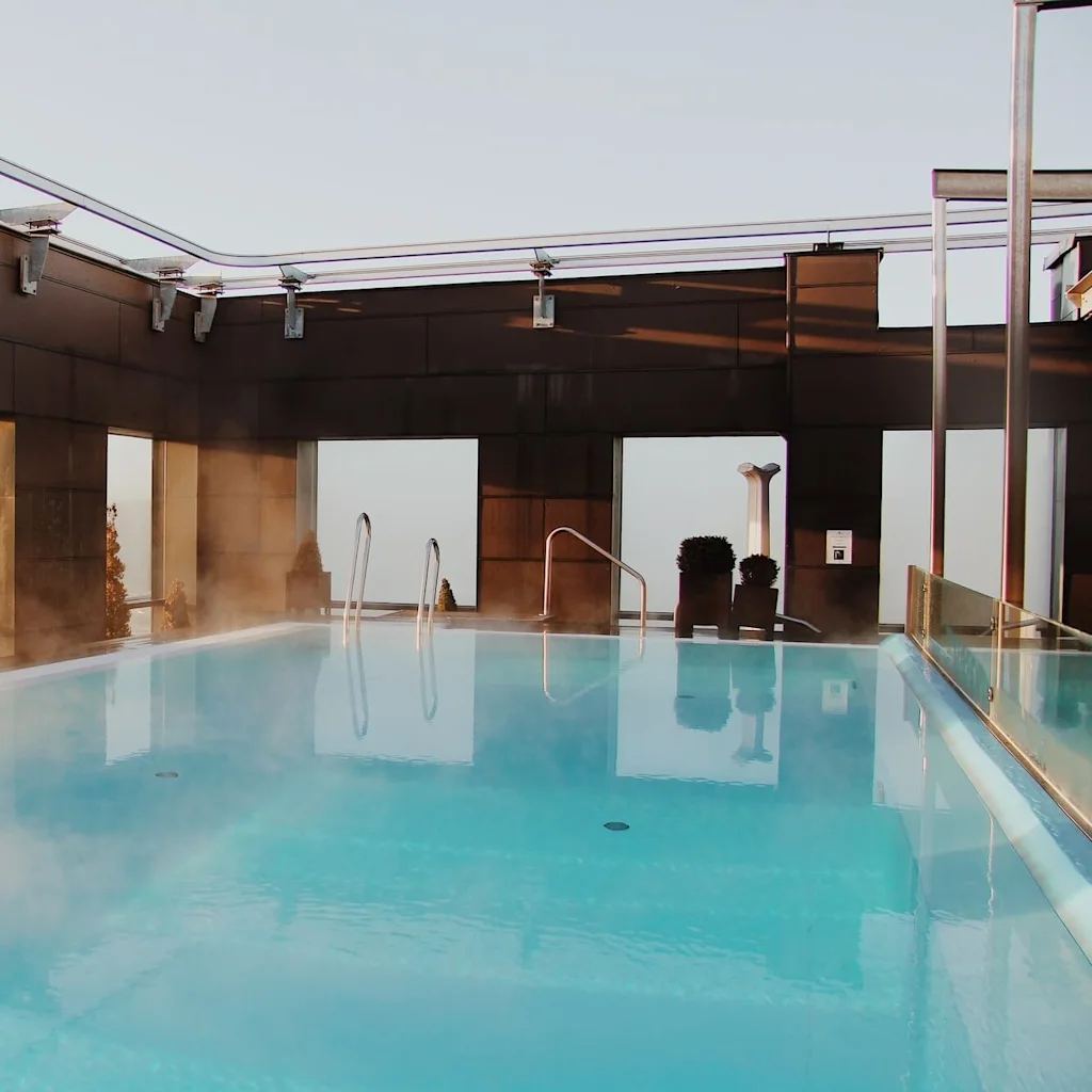 A steaming rooftop pool reflects morning light, surrounded by a modern metal and glass structure, offering a tranquil view of the sky above.