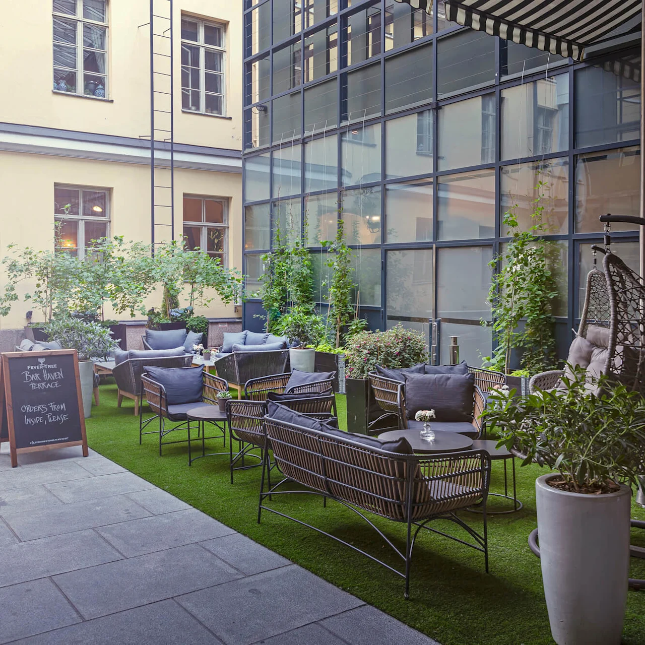 Cafe exterior with wicker chairs and tables set on artificial grass, surrounded by plants against a glass and yellow wall. A sign reads: "Take Away Terrasse Orders from Inside, Please”.
