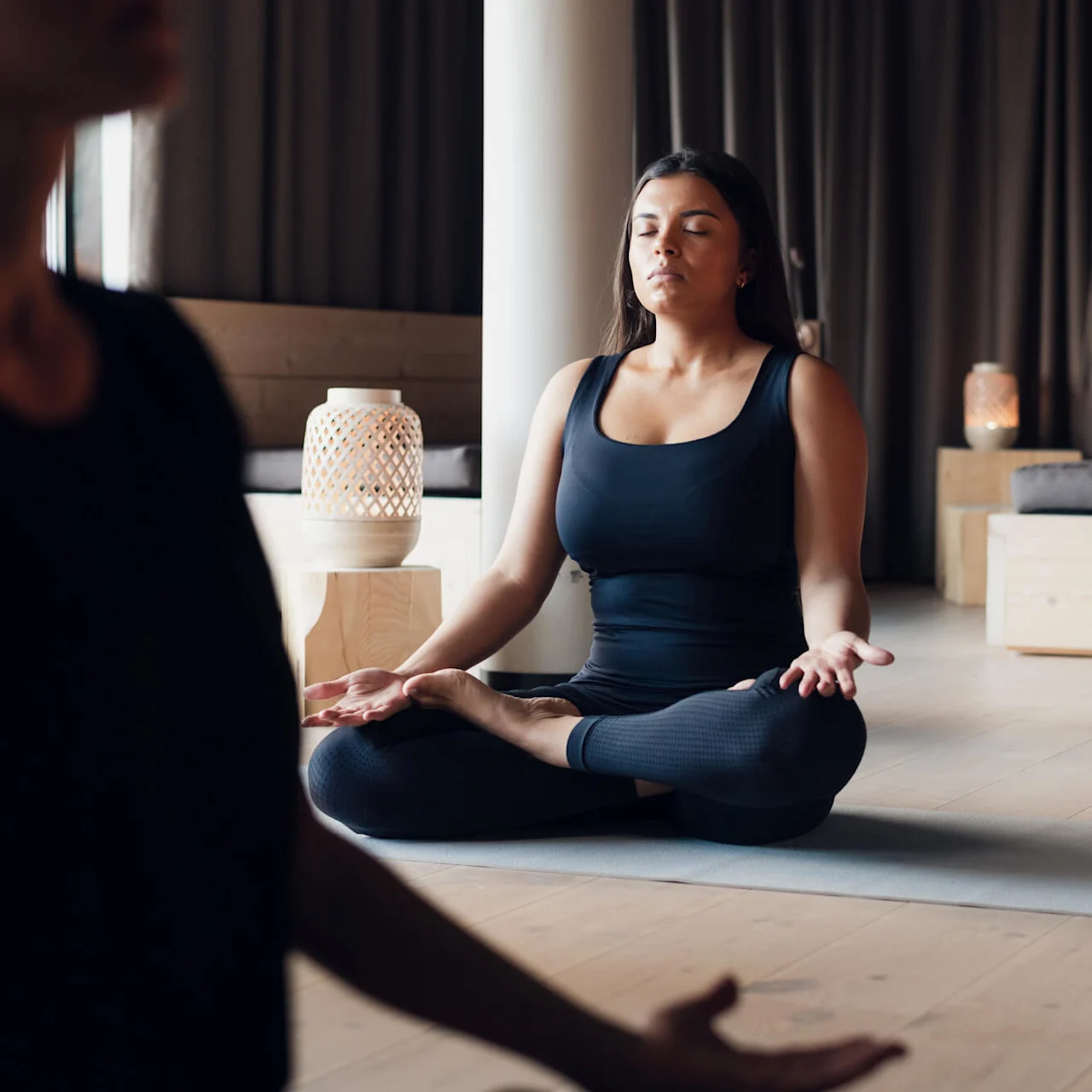Kvinna mediterar i korslagd ställning på en yogamatta med slutna ögon. Mjuk belysning, trägolv och dekorativa lampor skapar en fridfull atmosfär.