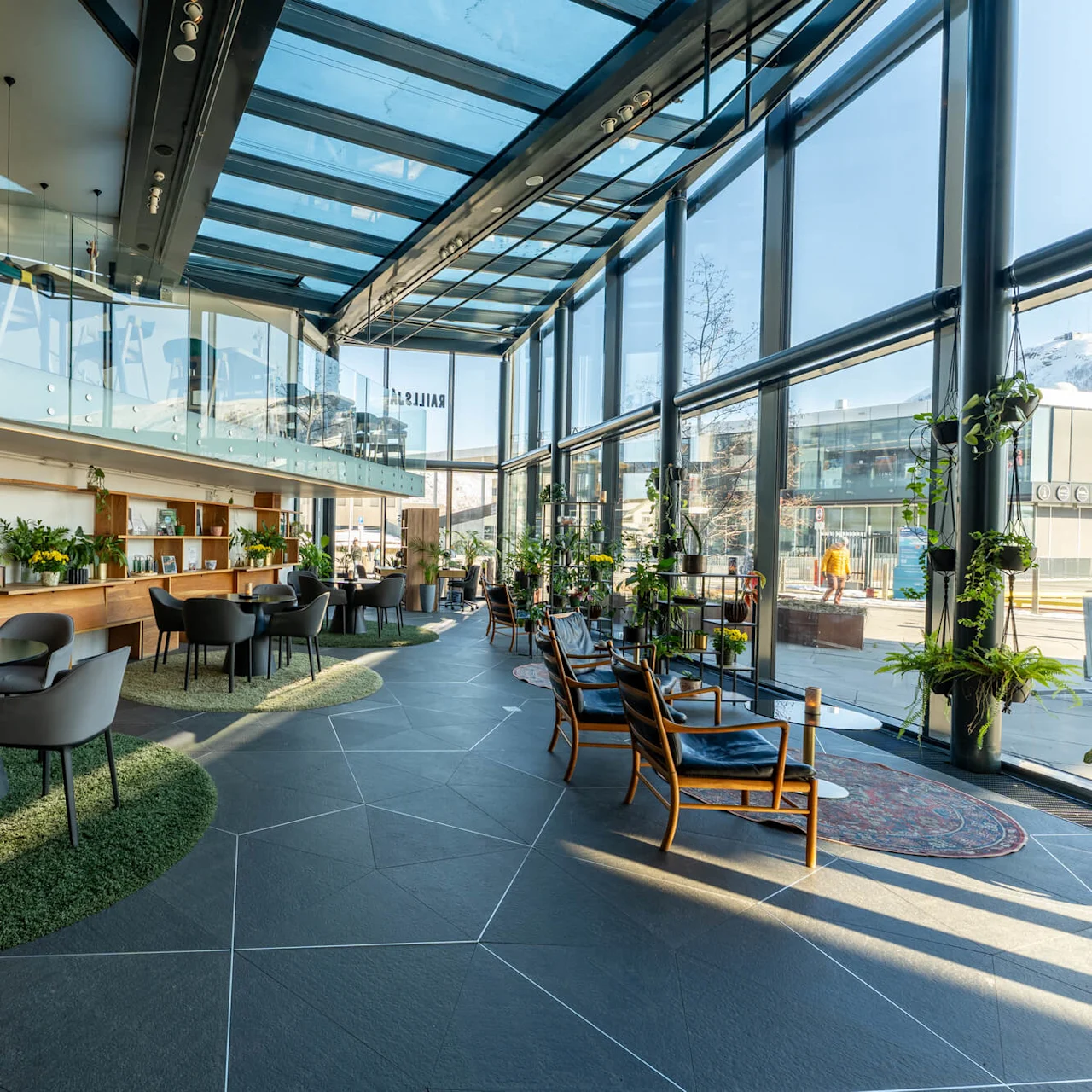 Borde og stole er arrangeret i en rummelig, moderne lounge med glastag og vægge. Bøger og planter pynter træhylderne og skaber en hyggelig og indbydende atmosfære.