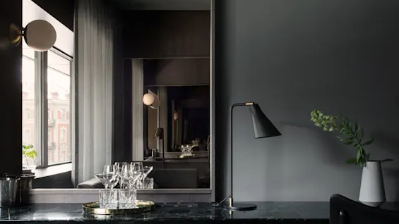 Et speil reflekterer et stilig rom med en mørk marmorbenk. Glass og et gullbrett står ved en svart lampe, nær et vindu med tynne gardiner.