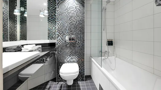 Et moderne badeværelse indeholder et væghængt toilet og et badekar, omgivet af hvide fliser. En mosaikflise fremhæver væggen; håndklæder og glas hviler på en bordplade.