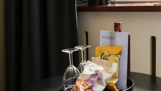 Et hotel minibar-arrangement med to vinglas, snacksposer og et MINIBAR-skilt på et lille bord ved et vindue.