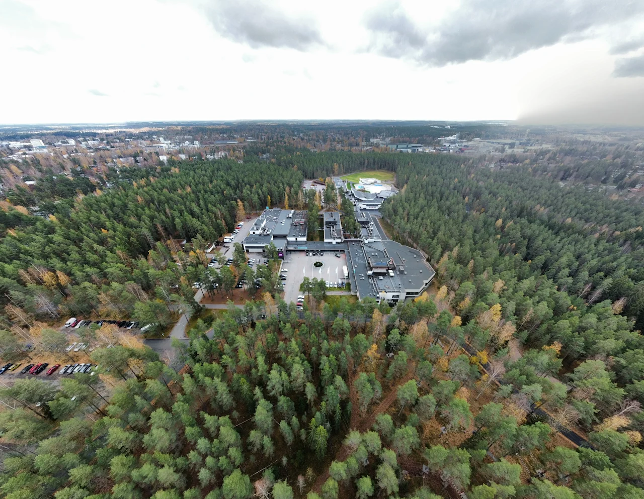 Suuri rakennuskompleksi on tiheän metsän ympäröimänä pilvisen taivaan alla, ja edustalla näkyy tie. Näkymä laajenee laajoihin metsämaisemiin.