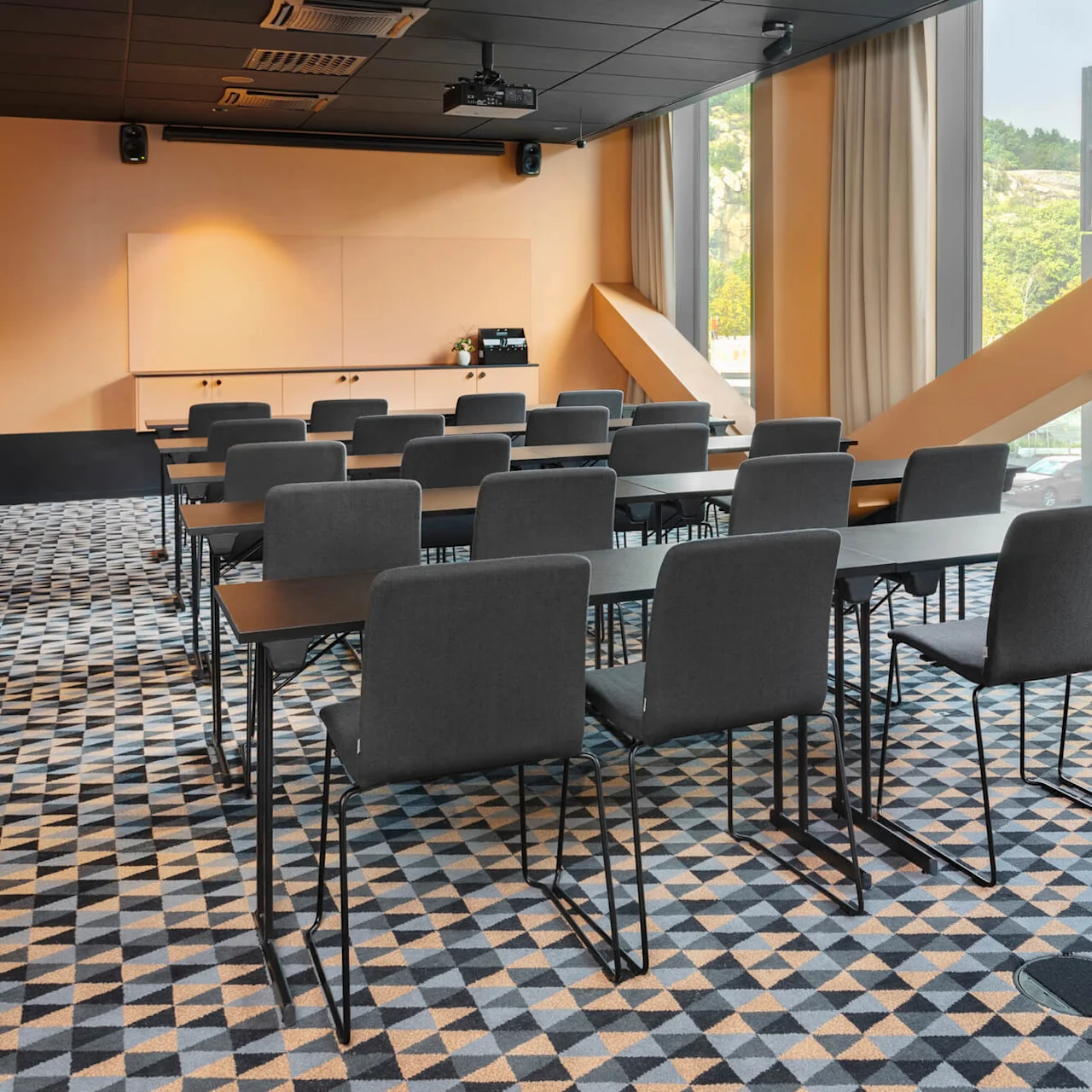 Rader med grå stolar vända mot en projektorduk i ett konferensrum. Stora fönster med gula balkar och geometriskt mönstrad matta förstärker den moderna miljön.