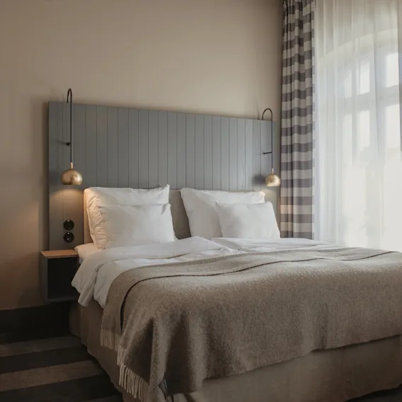 Et koselig hotellrom med en stor seng, hvite puter og et varmt teppe. Lys strømmer inn fra et vindu med rutete gardiner.