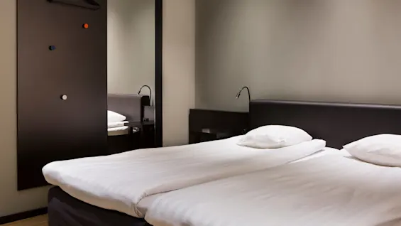 To enkeltsenge med hvidt sengetøj står side om side i et minimalistisk hotelværelse. Sorte sengelamper er fastgjort til sengegavlene. Et stort spejl og knager er på væggen.