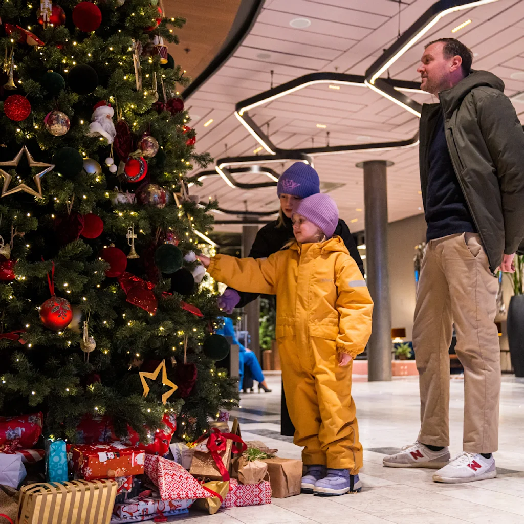 A decorated Christmas tree adorned with lights and ornaments stands on a tiled floor inside a building. A child in a yellow snowsuit and an adult observe it closely.