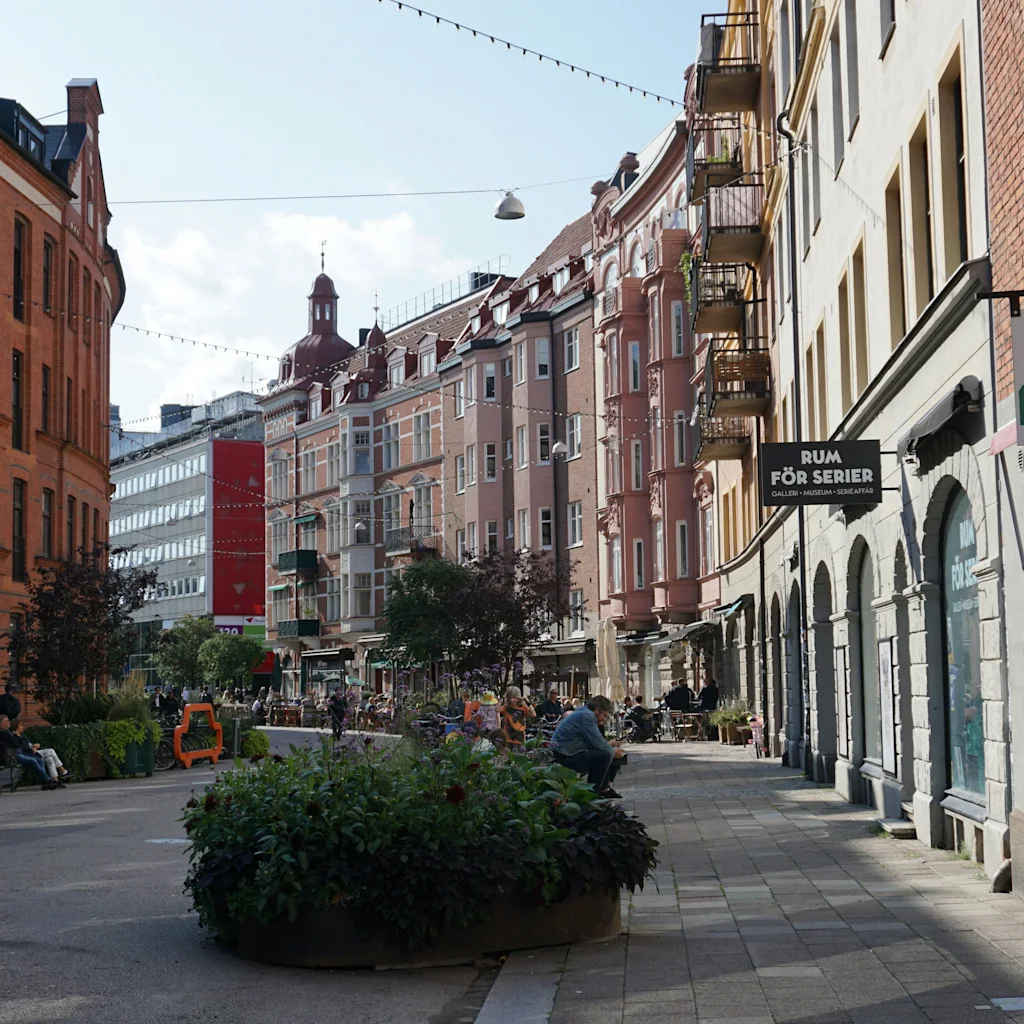 En travel gate i Malmö med historiske bygninger, trær og folk som nyter bymiljøet. Et skilt sier RUM FOR SERIER.