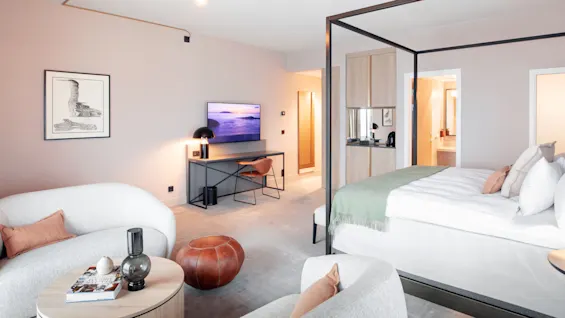 Et moderne hotellrom har en himmelseng, en koselig sittegruppe, en flatskjerm-TV montert på veggen og et skrivebord med en lampe, med et minimalistisk design.