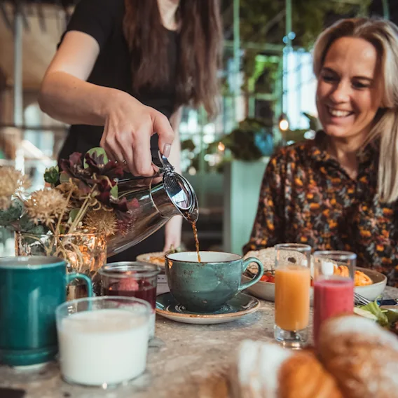 En servitør skjenker kaffe til en smilende gjest ved et frokostbord fylt med juice, smoothie og bakverk.