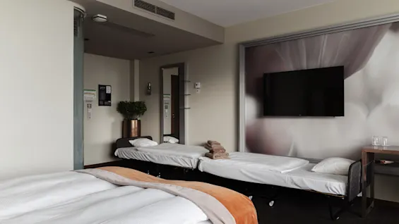 Ett hotellrum med en stor säng och två extrasängar, en väggmonterad TV och en växt, som erbjuder en bekväm vistelse.