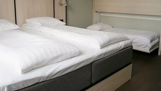 To enkeltsenger med hvitt sengetøy er pent plassert i et moderne rom, ved siden av en veggmontert utslagseng. Rommet har lyse trepaneler og en liten leselampe.