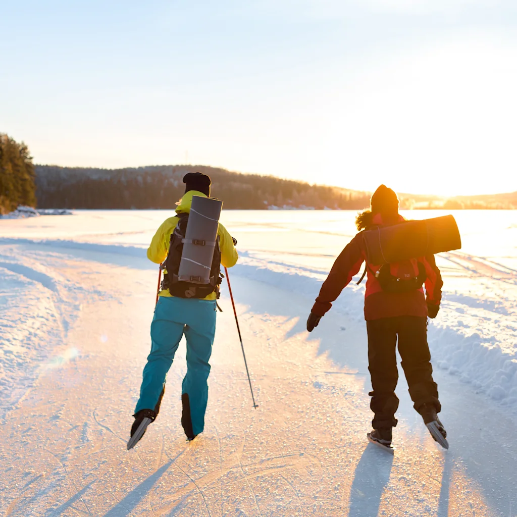Kaksi ihmistä luistelee jäätyneellä järvellä auringonnousun tai -laskun aikaan, lumipeitteisen luonnon ympäröimänä.