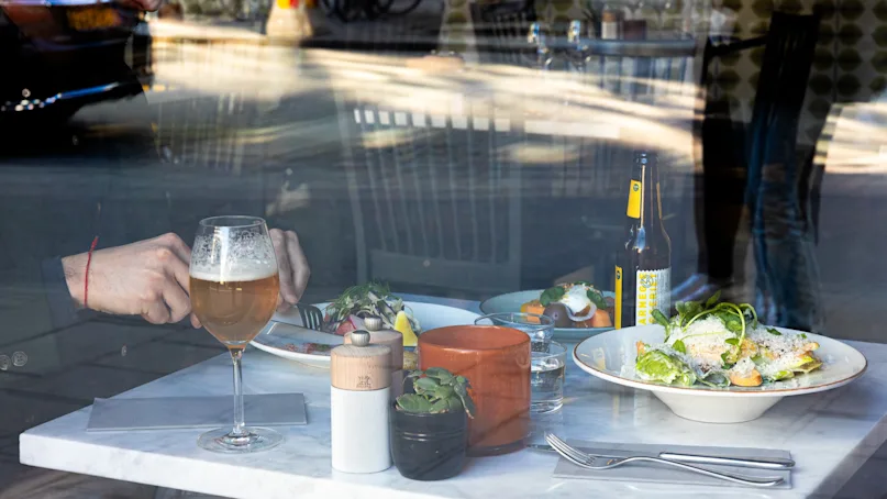 A person dines at a restaurant table, using utensils on a dish with vegetables, accompanied by a glass of beer, bottled drink, and potted plants, viewed through a window.