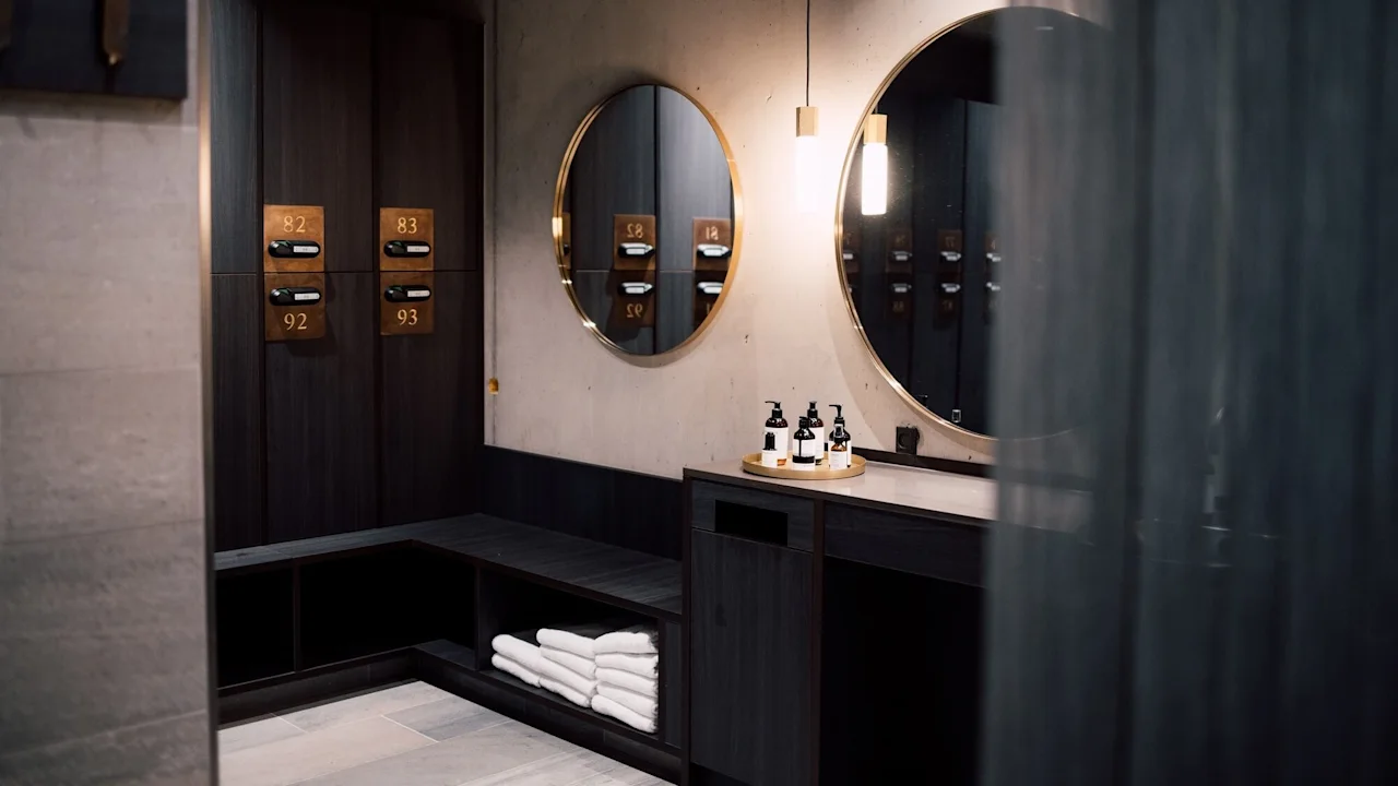 Lockers with numbers 82, 83, 92, 93 hang on a wall. Round mirrors reflect the room. Towels are neatly stacked below a counter with arranged soap dispensers in a modern restroom.