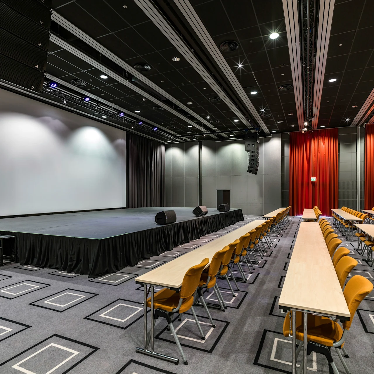 A large stage with microphones stands before long, rectangular tables with orange chairs in rows, set in a spacious conference room with patterned carpet and red curtains.