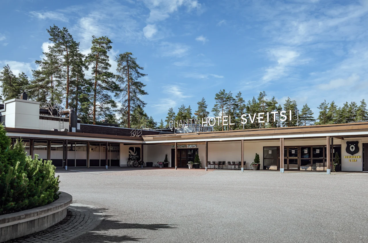 Suuri hotelli, jossa lukee "Quality Hotel Sveitsi" näkyvästi julkisivussa. Ympärillä laaja ajotie ja puita, taustalla kirkas taivas.