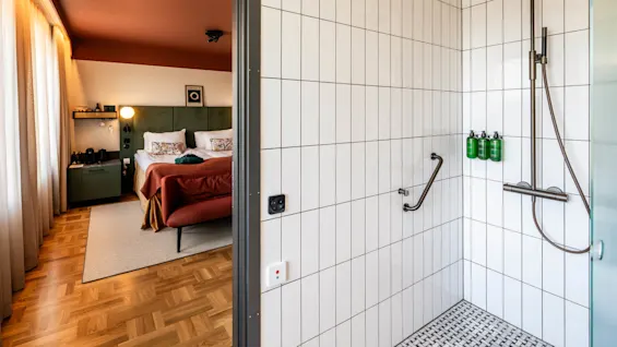Shower stall with white tiles and three green bottles on the wall. Beyond, a cozy bedroom with double bed, patterned pillows, and a side table, illuminated by natural light through curtains.