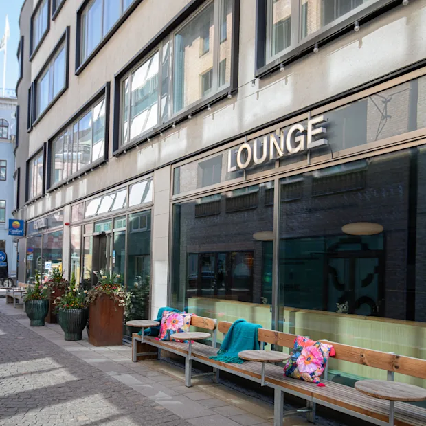 Et indbydende udendørs loungeområde med bænke og planter foran en bygning med store vinduer. Skiltet siger LOUNGE.