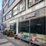 Et indbydende udendørs loungeområde med bænke og planter foran en bygning med store vinduer. Skiltet siger LOUNGE.