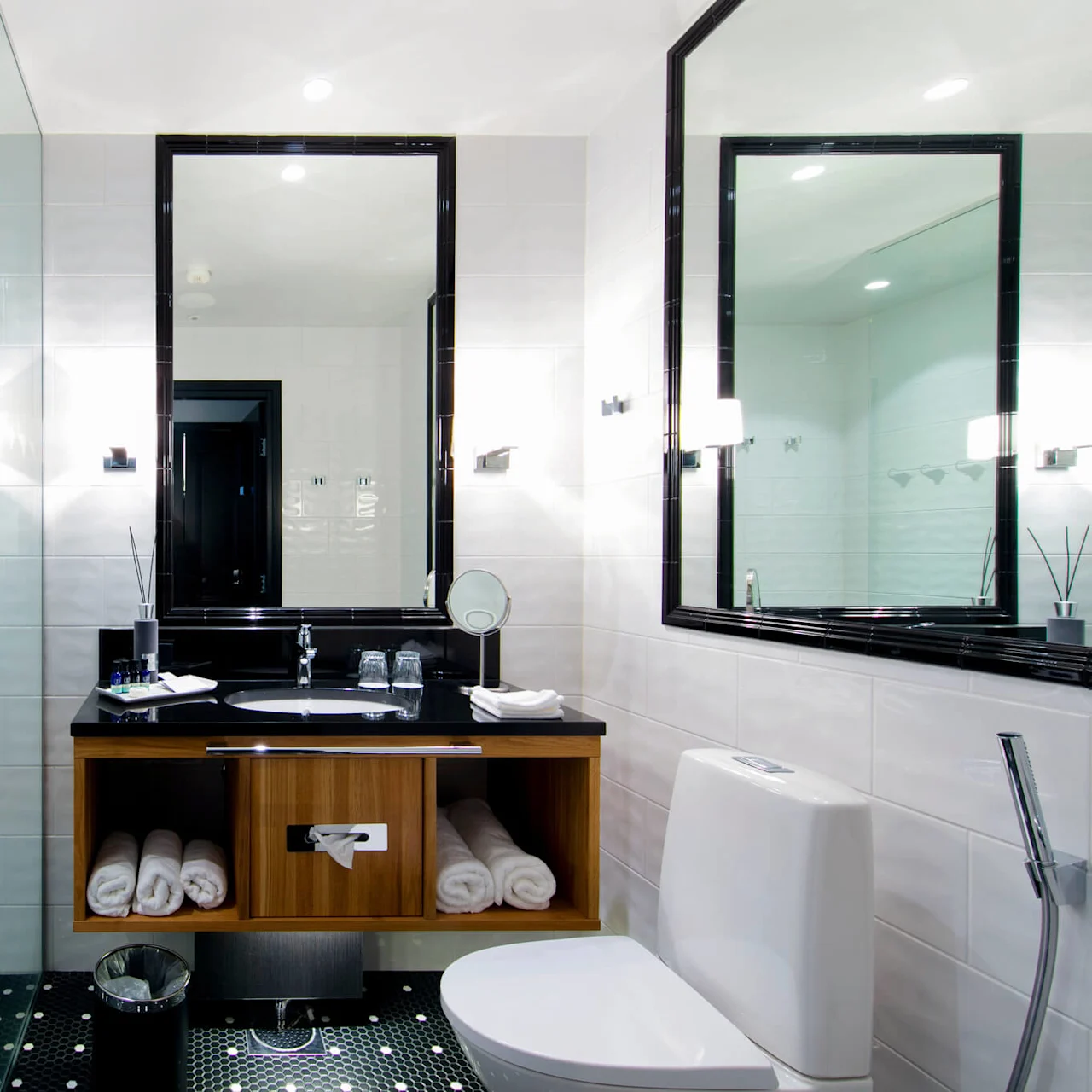 Et moderne bad med en brusekabine med glasvæg, en sort bordplade og en elegant toilet. Hvide fliser og spejle fremhæver det lyse, rene design.