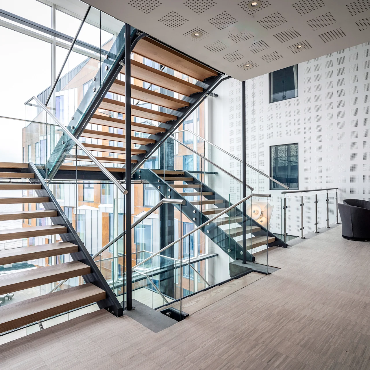 En elegant trappa i trä med glasräcken går upp och ner i ett ljust, modernt atrium, omgivet av stora glasfönster och prickiga väggar. Två svarta stolar och ett bord finns i närheten.