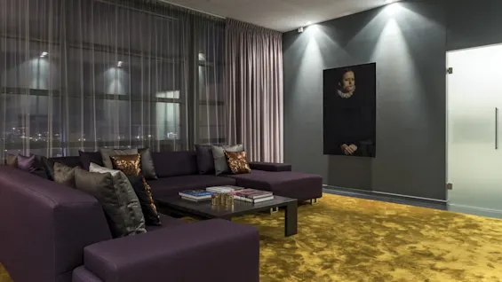 A purple sectional sofa sits in a modern living room, surrounded by a yellow rug and sheer curtains. A portrait hangs on a gray wall, illuminated by spotlights.