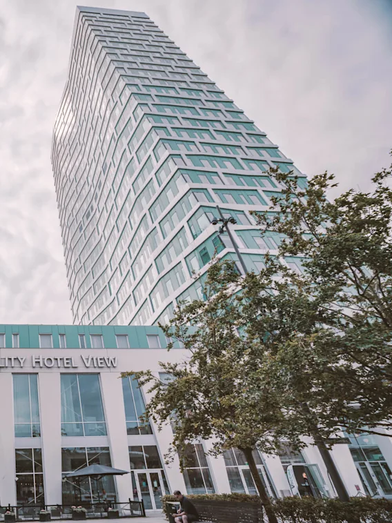 Two tall, modern buildings rise with a connecting structure labeled "Quality Hotel View." Trees and a cloudy sky surround the hotel entrance.