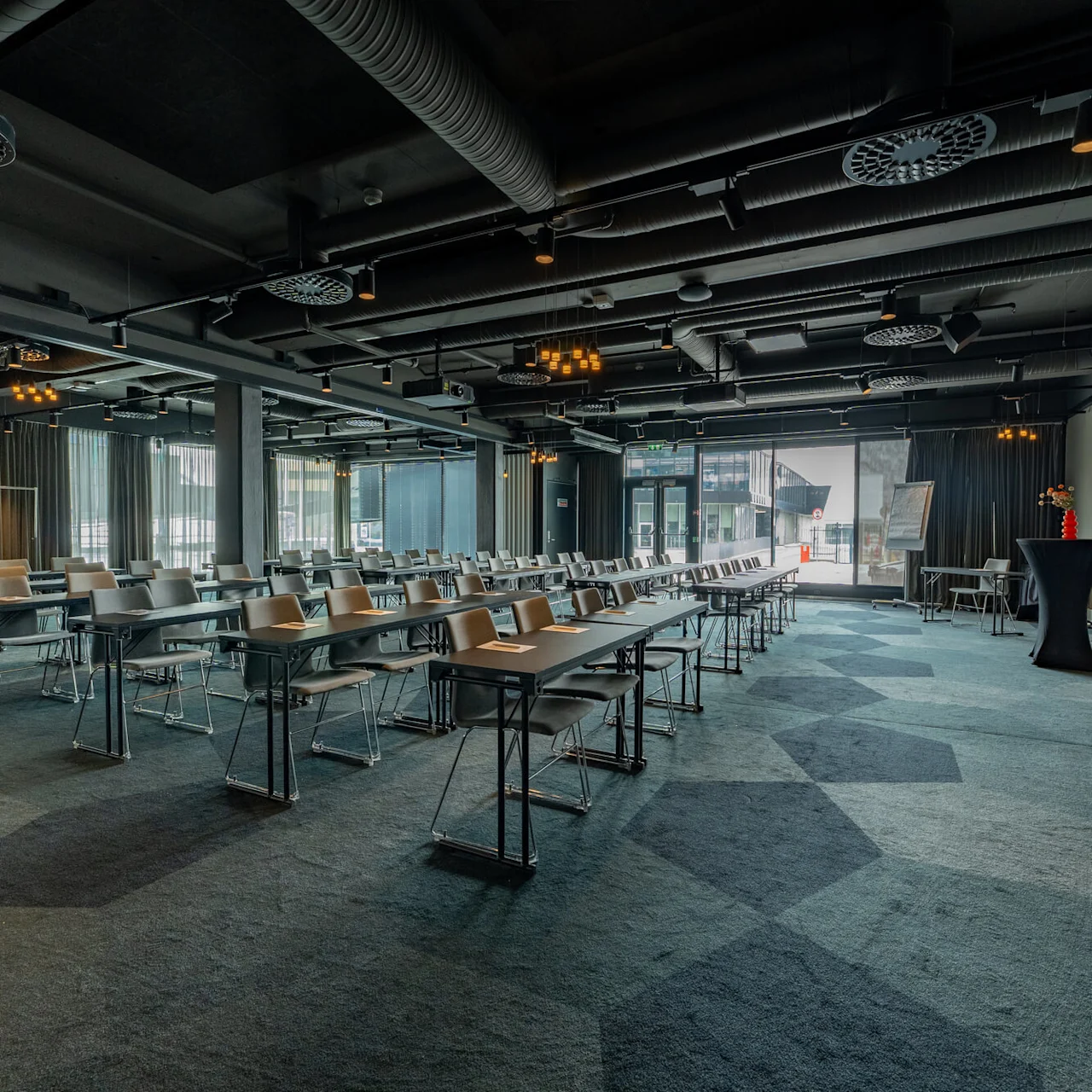 Konferencerum med rækker af tomme borde og stole, vendt mod en stor skærm, der viser "Clarion Hotel, CLARION THE EDGE." Rummet har moderne indretning og dæmpet belysning.