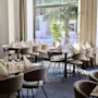 Tables are elegantly set with glasses and folded napkins in a modern restaurant. Soft chairs surround the tables, and large windows offer a view of trees outside.