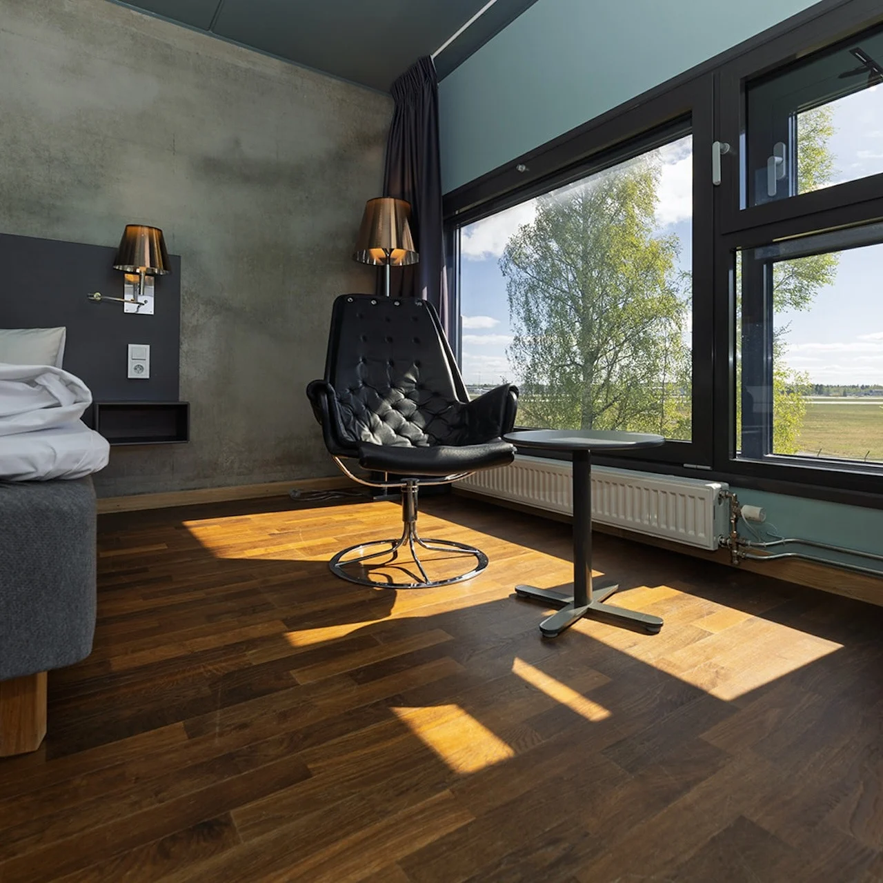 A modern hotel room with a stylish black chair facing a window, next to a bed with white sheets. Sunlight streams in, illuminating the wooden floor.