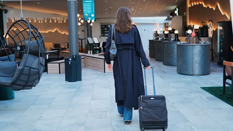 A person strolls through a modern hotel lobby with a suitcase, passing floating chairs and a reception desk beneath stylish lighting.