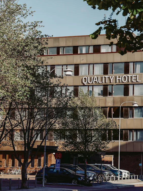 Kvalitetshotellskylt på en modern byggnad i flera våningar omgiven av träd och parkerade bilar. Träden skymmer delvis hotellutsikten och skapar en naturlig stadsbild.