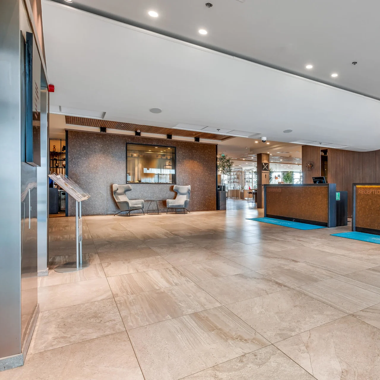 Modern hotel lobby with reception desks, tiled floor, seating area, and bright lighting.